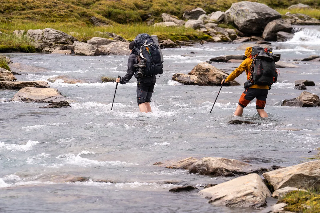 Two trekkers with large backpacks and trekking poles wading across a mountain stream