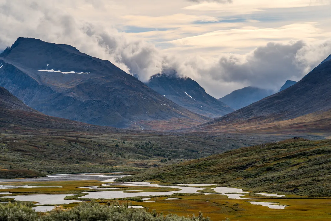 Mountain valley with a meandering river, dark slopes and low clouds, autumn tundra in the foreground